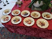 Set of nine Bavarian vintage plates laid out on a red tablecloth, showing various fruit designs including pears, peaches, grapes, and plums. Plates have scalloped edges and gold trim.