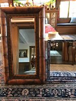 Large bamboo framed mirror with beveled glass standing upright beside a medium painted faux blue and silver framed mirror with hanging chains, both on an oriental-style rug in a living room setting.