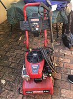 Full view of Craftsman gas powered pressure washer, red tubular frame, hose coiled on unit, pressure washer wand with trigger handle attached
