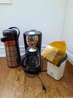 All three appliances together on wooden floor: coffee carafe, Oster coffee maker, West Bend popcorn maker.