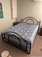Full view of black metal double bed with blue floral-patterned mattress in a carpeted room, showing headboard, footboard, and rails.