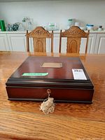 Closed wooden box with a metal plate on the lid and attached key in front, placed on a wooden table with chairs in background.