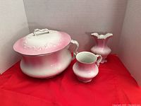 Set of three ceramic pieces: chamber pot with lid and handle, vase, cup, displayed on red cloth background and white backdrop