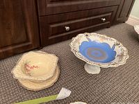 Pedestal fruit bowl with blue and gold detailing and two beige floral serving bowls on carpet in front of wooden cabinet.