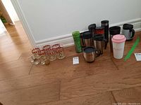 Overview of the lot showing all coffee travel mugs and glasses, arranged on wood floor.