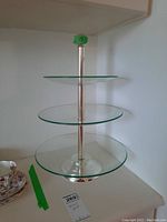 Front and side view of three-tiered dessert stand showing clear circular glass plates mounted on a silver-tone metal center pole with round base and ball finial top, approximately 19 inches tall.