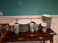 Wide view of multiple plates, bowls, and mugs stacked on wood cabinet, showing quantity and type of pieces.