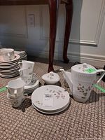 Full set of stacked tea cups, saucers, dessert plates, teapot, and sugar bowl arranged on carpet under wooden table legs.