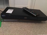 Front and top view of stacked Sony BDP-S350 Blu-ray DVD player with remote on top, and Philips DVP5960/37 DVD player beneath it on carpeted floor.