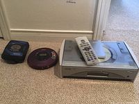Wide shot showing three devices on carpet: purple Sony Walkman portable CD player, black and blue Panasonic cassette player with radio, and JVC larger CD player with remote and manual on top of it.