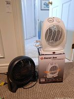 Photo of both fans together showing Comfort Zone heater fan in white on top of its box and black Honeywell fan on carpet below.