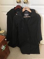 Two black ladies' jackets hanging on doors, showing front views. One is the Xterior lightweight spring jacket with hood and button front (size S). The other is the Kenneth Cole Reaction winter coat, 80% wool, larger size XL, with buttons and hood.