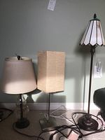 Photo showing three floor lamps lined up against a wall, varying heights, one with stained glass shade, one with square beige shade, and one with beige fabric shade on glass base.