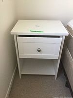 White melamine nightstand against wall next to bed, drawer closed, showing round metal knob and clean flat top surface