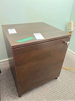 Front view of one nightstand showing wood veneer finish, cabinet door with round brushed metal knob, flat top surface, and small legs.