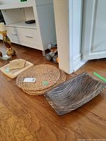 Photo showing two round wicker-style woven placemats, one square wooden tray with geometric pattern, and one rectangular wooden tray with carved wave pattern arranged on wooden floor near white cabinet.