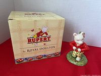 Figurine of Rupert Bear in red jacket and yellow checkered pants, standing on green base with The Little Sea Sprite figure on his back, displayed next to original box with Rupert branding.