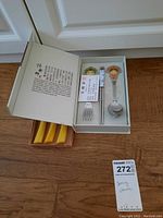 Boxed set of serving utensils including fork, spoon and chopsticks inside an open box with printed text on lid and paper certificate.