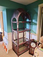 Full view of the tall wicker shelf with glass shelves, showing the overall design and structure in a corner against a teal wall with a clock beside it.
