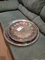 Two old English silver plate trays stacked on a gray couch. Smaller ornate tray with floral engraving and scalloped edges sits on top of a larger hammered silver plate tray.