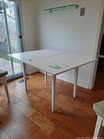 View of the convertible kitchen table in open position showing white melamine top with visible marks and sturdy white wooden legs on wood flooring near sliding glass door.