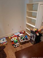 Children's toys scattered on hardwood floor near shelving unit, including cars, trucks, plane, and baskets of toys