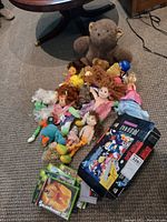 Pile of soft toys on carpet including large teddy bear, rag dolls, vinyl baby dolls and small plush figures
