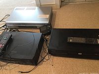 Four electronic devices on carpet: black Toshiba DVD player with remote, black Sharp VCR, black Sony DVD and VHS player, and a portable Sirius radio device.