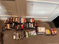 Overview of VHS tapes, DVDs, and CDs arranged on the floor including varied titles and formats.