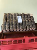 Ten leather bound antique books arranged upright on a wood book stand, showing detailed gold embossing on the spine with some wear and chips on top edges.