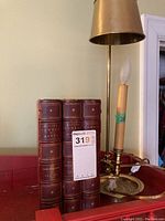 Three leather-bound volumes of The Divine Comedy placed upright, titles visible on spines along with a brass lamp with candle sleeve marked '20'.