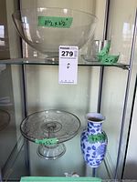 Clear glass large bowl and small glass bowl with measurements visible on tape. Blue and white floral vase beside cake plate.