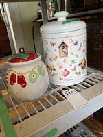 Two ceramic cookie jars displayed on a shelf: one smaller Christmas mittens design, one larger garden motif signed Maryjolein Boston