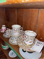 Photo showing three cups and saucers with floral and gold decorations on a wooden shelf.