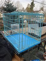 Angle view of small blue wire dog crate showing front and side, placed outdoors on a table with background of trees and houses.