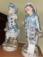Full view of the pair of porcelain figurines, boy and girl, standing on bases with blue and white themed clothing.