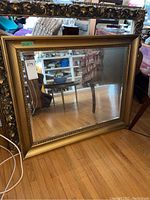 Full view of rectangular gold framed mirror resting on wooden floor with surrounding household items.