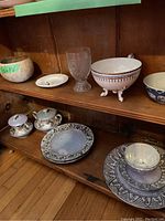 Full lot items arranged on wooden shelving, shows metallic lusterware cream and sugar, footed bowl, glass goblet, small ashtray and multiple plates.