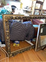 Full view of the large carved wood frame leaning on a chair in a cluttered room, showing the overall size and finish.
