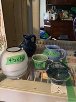 Four pottery items on reflective surface with size labels: white and pale blue vase (4.5 x 4.5 inches), green bowl (4 x 2 inches), pale purple creamer (3.5 x 5 inches), dark green honey pot.