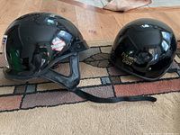 Photo shows two glossy black motorcycle helmets placed on a wooden floor, one Harley-Davidson open-face helmet with chin strap on the left and one Fulmer Trooper DOT half-shell helmet on the right. Both helmets have logos on the side and are visibly used but intact.