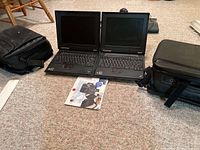 Photo showing two IBM ThinkPad laptops opened side by side on carpet with two black carrying cases on either side and a ThinkPad manual on the floor in front.