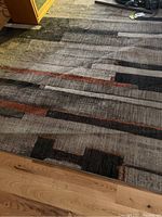 Photo shows rectangular 8x10 area rug with geometric blocks and linear patterns in gray, black, and rust colors. Rug laid on wood floor. Rug appears worn and needs cleaning.