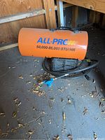 Orange All Pro forced air heater with propane hose and blue propane tank connector sitting on ground in wooden shed.