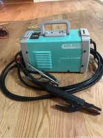 Portable teal and gray YESWELDER ARC-205DS welder on wood floor with attached cables and package of electrodes