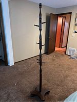 Full view of tall wooden coat rack with multiple hooks and tripod base in a room with carpet flooring.