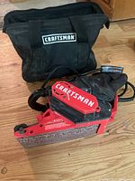 Photo showing the Craftsman red and black belt sander with dust bag attached next to a black fabric carrying case with the Craftsman logo.