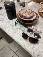 Black Cuisinart coffee grinder, four ceramic bowls stacked with various decorative patterns, and three pairs of sunglasses in different styles and colors on a marble countertop.
