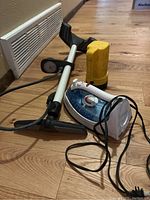 Photo shows yellow flashlight, white and gray bicycle pump with pressure gauge, and black and white iron with cord in a row on wooden floor.