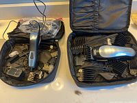 Photo showing two black carrying cases containing hair cutting tools, comb attachments, and power cords. One trimmer is labeled Wahl, other is unidentified but similar style.